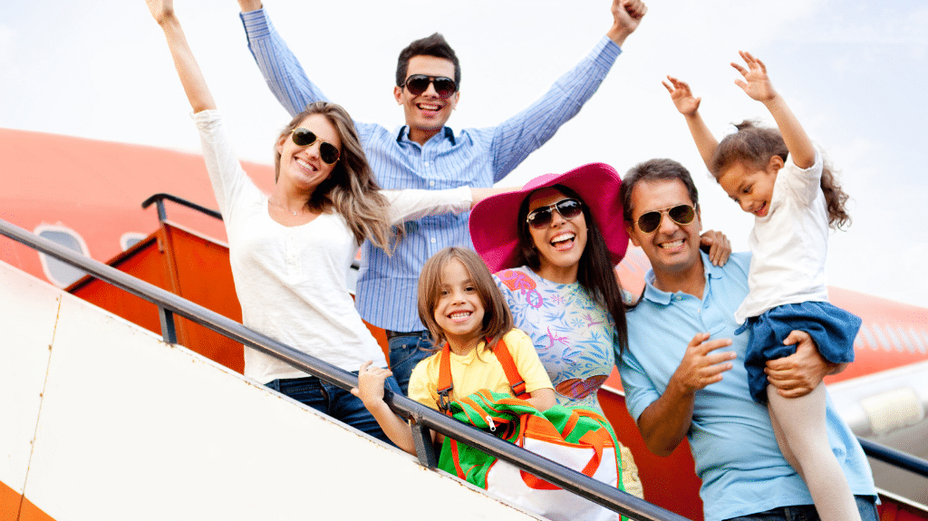 Family traveling with children pose outside of an airplane with arms up in excitement.